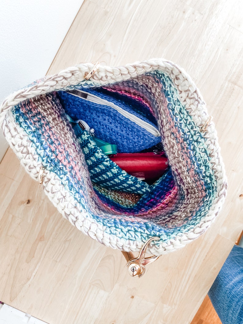 Pode incluir: Uma bolsa de ombro de croch&ecirc; com um padr&atilde;o listrado em tons de rosa, azul e verde. A bolsa tem al&ccedil;as douradas e est&aacute; cheia de v&aacute;rios itens, incluindo uma bolsa azul, uma bolsa vermelha e uma bolsa verde.