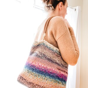 Pode incluir: Uma bolsa tote de croch&ecirc; listrada multicolorida com al&ccedil;as de couro marrom. A bolsa &eacute; feita com uma variedade de tons de fio marrom, azul, rosa e verde.