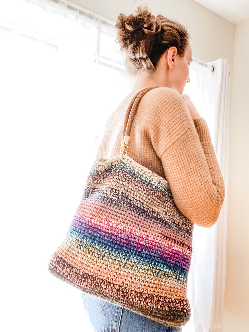 Pode incluir: Uma bolsa tote de croch&ecirc; listrada multicolorida com al&ccedil;as de couro marrom. A bolsa &eacute; feita com uma variedade de tons de fio marrom, azul, rosa e verde.