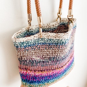 Pode incluir: Uma bolsa tote de croch&ecirc; listrada multicolorida com al&ccedil;as de couro marrom e ferragens douradas.