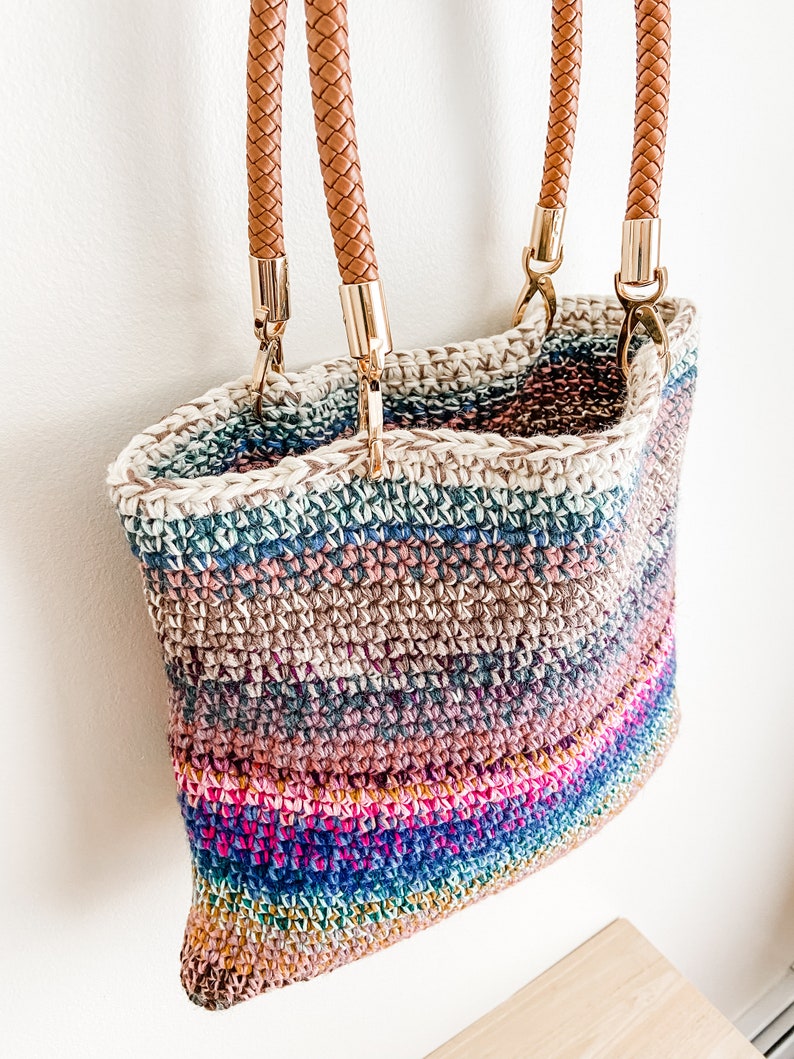 Pode incluir: Uma bolsa tote de croch&ecirc; listrada multicolorida com al&ccedil;as de couro marrom e ferragens douradas.