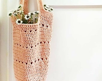 May include: A pale pink crocheted tote bag with a delicate openwork pattern hangs on a white door. The bag is filled with a bouquet of white flowers.