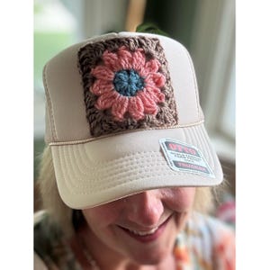 May include: A beige trucker hat featuring a square crochet patch on the front panel. The patch displays a flower design with a blue center, pink petals, and a brown border. A sticker on the brim reads OTTO WORLD FAMOUS FOAM FRONT OSFM TRUCKER.