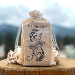 Może przedstawiać: Biała torba z pł&oacute;tna z sznurkiem do ściągania. Na torbie nadrukowany jest napis "NEW MOON TAROT". Torba zawiera czarno-biały rysunek dw&oacute;ch twarzy z gałązką liści między nimi.