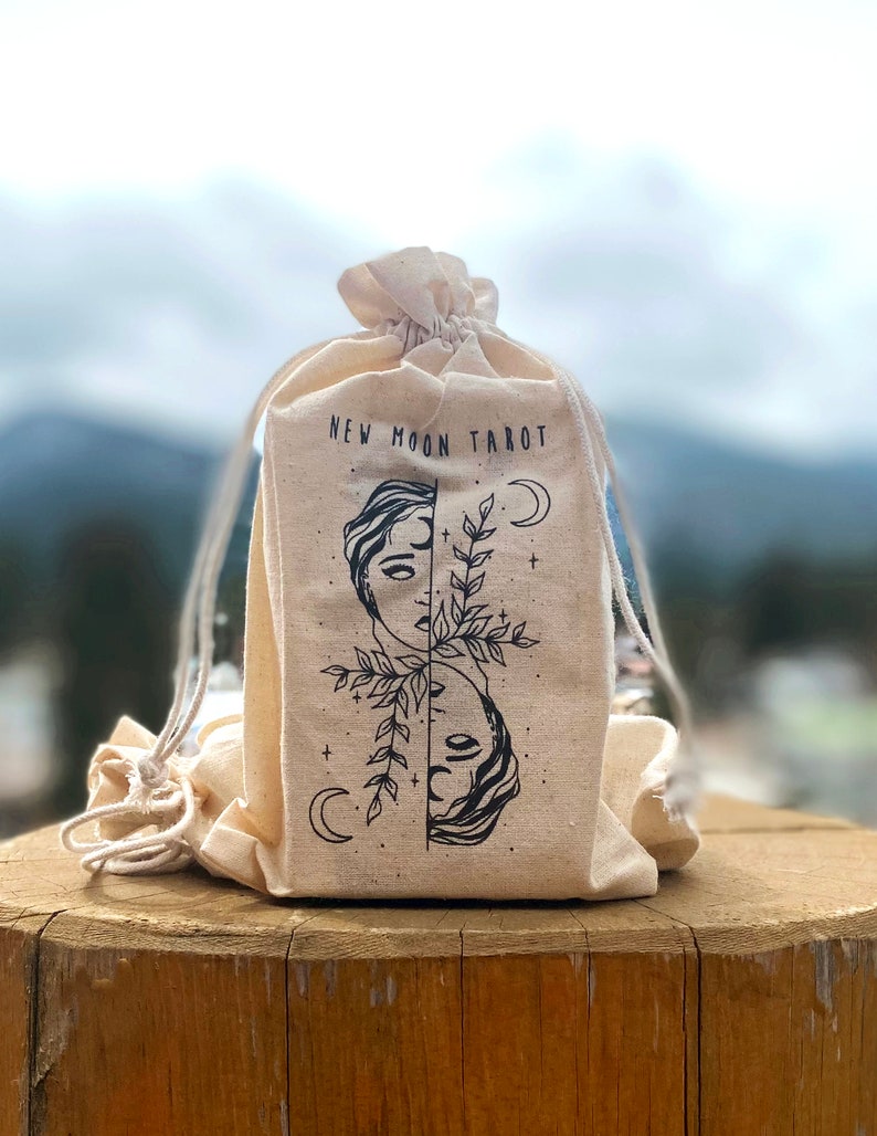 Może przedstawiać: Biała torba z pł&oacute;tna z sznurkiem do ściągania. Na torbie nadrukowany jest napis "NEW MOON TAROT". Torba zawiera czarno-biały rysunek dw&oacute;ch twarzy z gałązką liści między nimi.