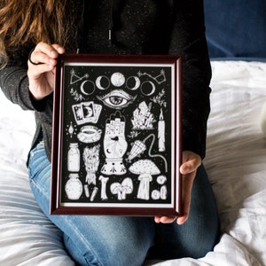 May include: A black and white illustration print featuring a hand with an eye, moons, crystals, mushrooms, and other mystical symbols. The print is framed in a dark wood frame.