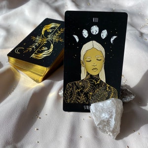 Może przedstawiać: Czarna i złota karta tarota z numerem VIII i słowem "Siła" na dole. Karta przedstawia kobietę z zamkniętymi oczami i kwiatowym wzorem na klatce piersiowej. Tło jest czarne z pięcioma białymi księżycami.