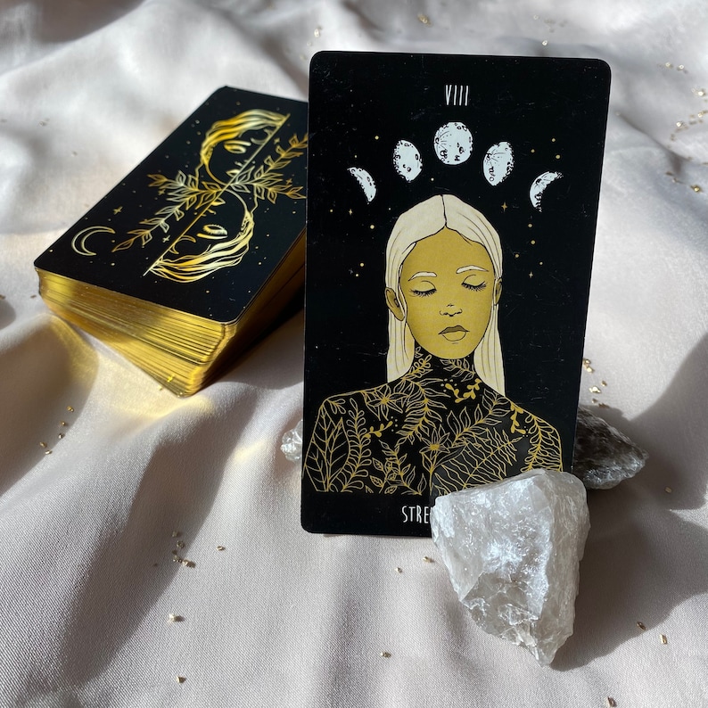 Może przedstawiać: Czarna i złota karta tarota z numerem VIII i słowem "Siła" na dole. Karta przedstawia kobietę z zamkniętymi oczami i kwiatowym wzorem na klatce piersiowej. Tło jest czarne z pięcioma białymi księżycami.