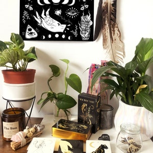 May include: A black and white illustration of a hand with a ring, a crescent moon, an eye, and a smudge stick. The illustration is on a black background with white stars. The illustration is framed and hanging on a white wall. There are tarot cards, a candle, and other items on a wooden table in front of the illustration.