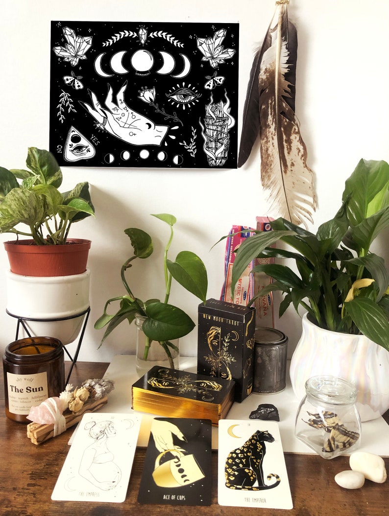 May include: A black and white illustration of a hand with a ring, a crescent moon, an eye, and a smudge stick. The illustration is on a black background with white stars. The illustration is framed and hanging on a white wall. There are tarot cards, a candle, and other items on a wooden table in front of the illustration.