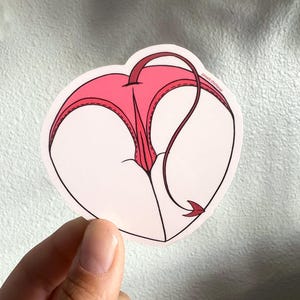 May include: A pink heart-shaped sticker with a red devil tail. The sticker has a stitched design and the text "@meili.thelover" is printed on the tail.