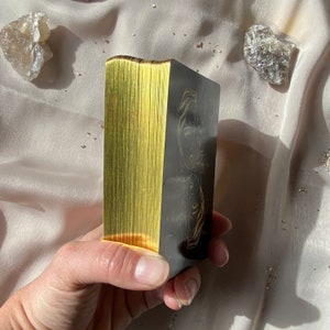 Może przedstawiać: Talia kart tarota z pozłacanymi krawędziami. Karty są ułożone w stos i przedstawiają czarny i złoty wz&oacute;r z kobiecą twarzą z przodu.