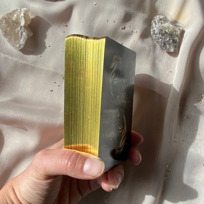 Może przedstawiać: Talia kart tarota z pozłacanymi krawędziami. Karty są ułożone w stos i przedstawiają czarny i złoty wz&oacute;r z kobiecą twarzą z przodu.