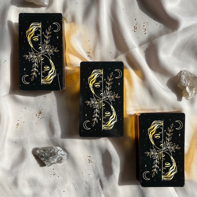 Może przedstawiać: Trzy czarne karty z wzorami w złotej folii przedstawiającymi twarz kobiety podzieloną na p&oacute;ł z gałązką liściastą i p&oacute;łksiężycami. Karty znajdują się na białym tle z tkaniny.