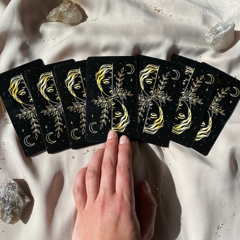 Może przedstawiać: Siedem czarnych kart z wzorami z złotej folii przedstawiającymi twarz kobiety, liście i rosnące księżyce. Karty są rozłożone na beżowej powierzchni tkaniny.
