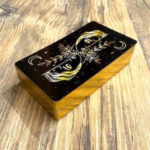 Może przedstawiać: Talia kart tarota z złotymi akcentami. Karty mają czarne tło z złotą ilustracją dw&oacute;ch twarzy i rosnącego księżyca.