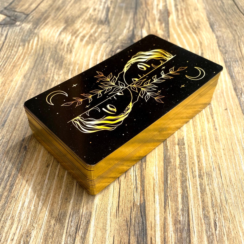 Może przedstawiać: Talia kart tarota z złotymi akcentami. Karty mają czarne tło z złotą ilustracją dw&oacute;ch twarzy i rosnącego księżyca.