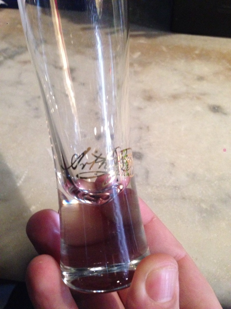 Op de afbeelding: Een helder glazen shotglas met een gouden inscriptie aan de zijkant. De inscriptie is moeilijk te lezen.
