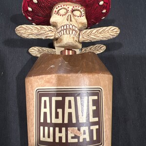 May include: A wooden beer tap handle with a red, white, and brown design. The handle features a skull wearing a sombrero and the words "Agave Wheat".