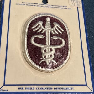 Puede incluir: Un parche bordado blanco y marrón con un símbolo médico, un caduceo, en una tarjeta blanca con el texto "N.S. MEYER, INC." y "OUR SHIELD GUARANTEES DEPENDABILITY".