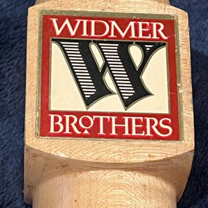 Puede incluir: Un tirador de cerveza de madera con una etiqueta roja y dorada que dice "Widmer Brothers" y presenta una "W" estilizada en blanco y negro.