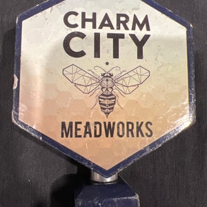 May include: A hexagonal tap handle with the words "CHARM CITY MEADWORKS" in black lettering. A geometric bee graphic is centered below the words. The handle has a silver metal base and a dark blue stem.