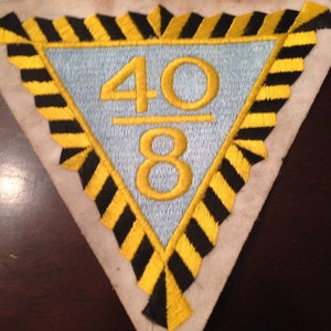 Puede incluir: Un parche triangular con un borde rayado negro y amarillo. El centro del parche es azul claro con los números 40 sobre 8 bordados en amarillo.
