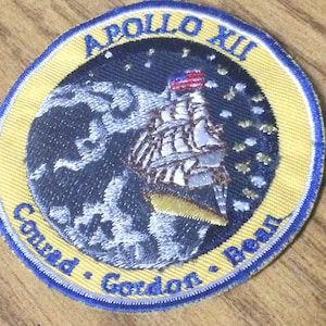 Może przedstawiać: Naszywka haftowana na żółto i niebiesko z tekstem "Apollo XII" i "Conrad, Gordon, Bean". Naszywka przedstawia stylizowane zdjęcie Ziemi z rakietą i flagą amerykańską.