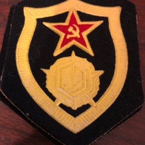 May include: A black felt patch with a gold embroidered shield. The shield features a red star with a hammer and sickle inside, and a gold circular emblem with a stylized design.