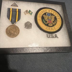 May include: A black framed display case containing a gold medal, a gold pin, two small American flags, and a gold embroidered patch with the words "United States Army" and "USA".
