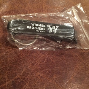 Puede incluir: Abrebotellas de llavero negro con un logotipo blanco que dice "Widmer Brothers Brewing" y un logotipo "W" blanco.