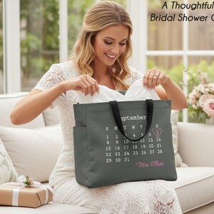 May include: A dark gray tote bag featuring a white calendar print for the month of September. The design includes a pink heart around the number 7 and the text Mrs. Elliott in pink script. The bag has black handles and side pockets.