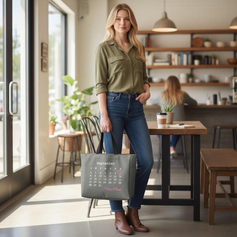 May include: A grey tote bag featuring a printed September calendar with a small pink heart icon on one date and the name Mrs. Elliott in pink script text. The bag has black handles and is held by a person wearing a green button-down shirt and blue jeans.