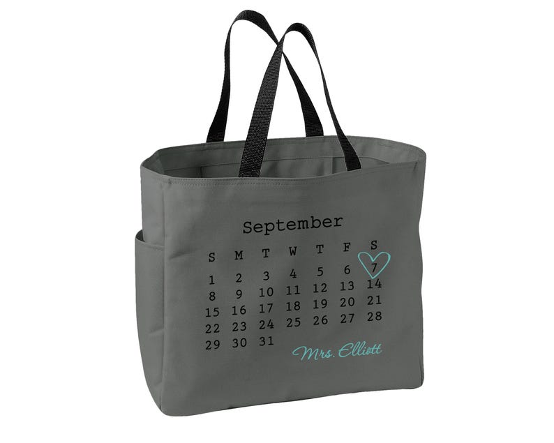 May include: A gray tote bag with black handles featuring a printed September calendar. The number 7 is circled with a light blue heart, and the text Mrs. Elliott is printed in light blue script below the calendar dates on the front of the bag.