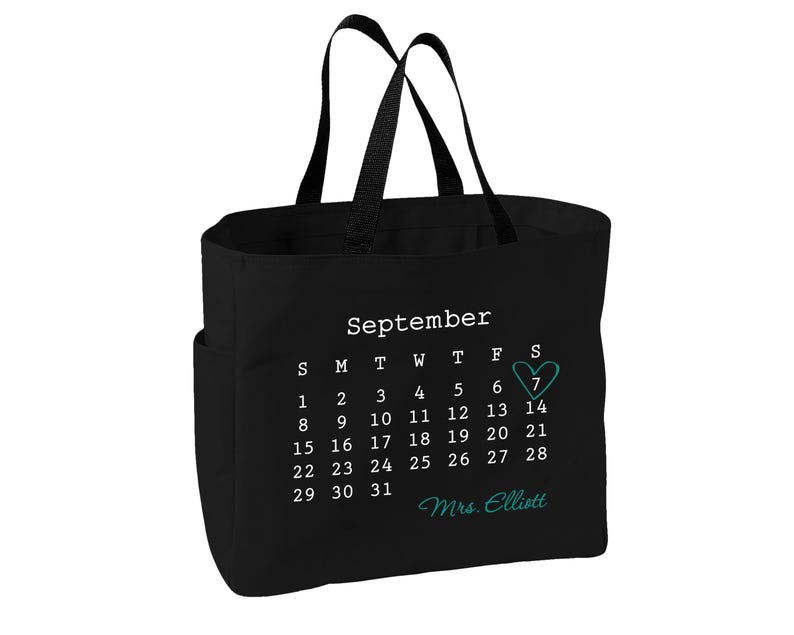 May include: A black tote bag featuring a white printed calendar for the month of September. The number 7 is highlighted with a teal heart outline. Below the calendar, the text Mrs. Elliott is printed in a teal cursive script font.