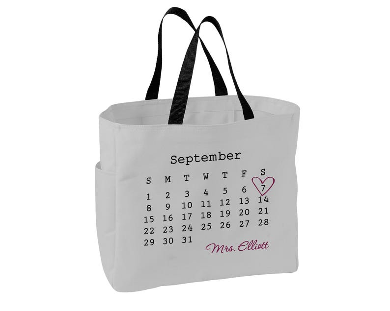 May include: A white canvas tote bag with black handles featuring a printed September calendar. The number 7 is circled with a pink heart, and the name Mrs. Elliott is printed in pink script below the calendar dates on the front of the bag.