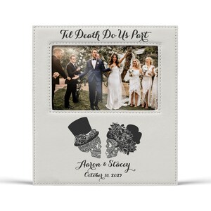 Op de afbeelding: Een witte fotolijst met de tekst "Til Death Do Us Part" bovenaan. De lijst bevat een trouwfoto en heeft schedelillustraties met de namen "Aaron & Stacey" en de datum "October 31, 2027".