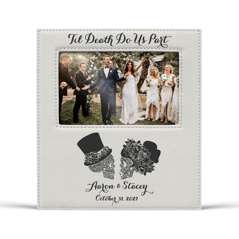 Op de afbeelding: Een witte fotolijst met de tekst "Til Death Do Us Part" bovenaan. De lijst bevat een trouwfoto en heeft schedelillustraties met de namen "Aaron & Stacey" en de datum "October 31, 2027".