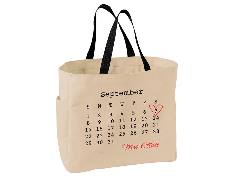 May include: A beige canvas tote bag with black handles featuring a printed September calendar. The number 7 is circled with a red heart, and the name Mrs. Elliott is printed in red script below the calendar dates on the front of the bag.