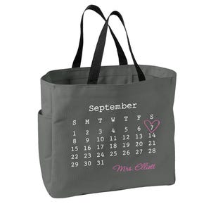 May include: A dark gray tote bag with black handles featuring a white printed September calendar. The number 7 is circled by a pink heart, and the text Mrs. Elliott is printed in pink script below the calendar dates on the front of the bag.