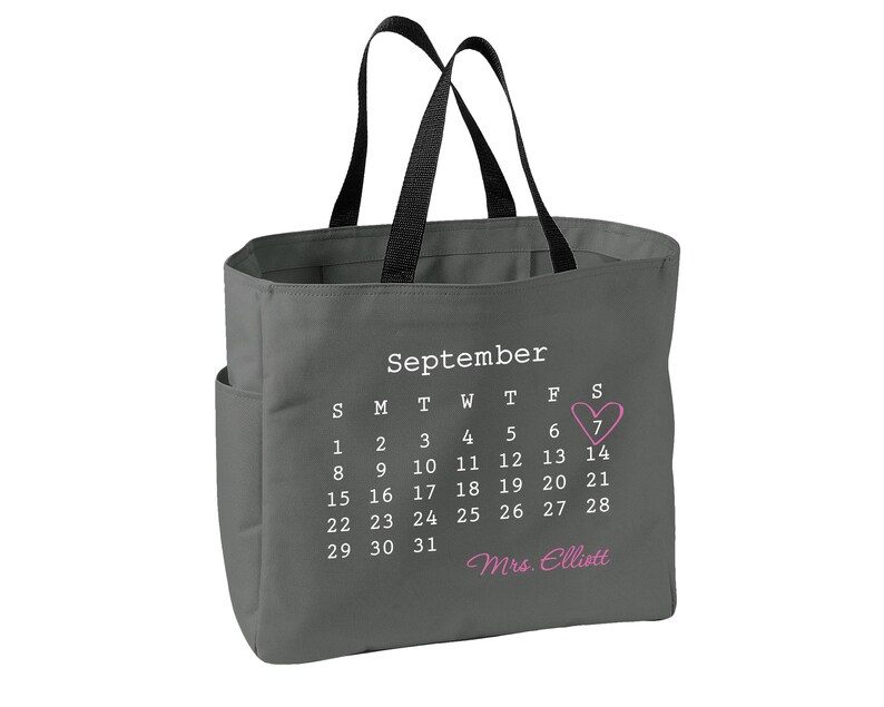 May include: A dark gray tote bag with black handles featuring a white printed September calendar. The number 7 is circled by a pink heart, and the text Mrs. Elliott is printed in pink script below the calendar dates on the front of the bag.