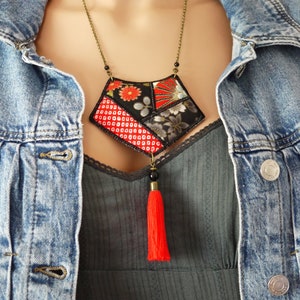 Peut inclure: Un collier original avec un pendentif géométrique en tissu à motifs rouges, noirs et floraux. Le collier a une longue chaîne avec de petites perles et un pompon rouge vif. Le mannequin porte une veste en jean.