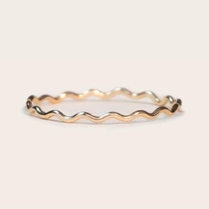 May include: A delicate gold ring with a wavy, textured design. The ring's surface reflects light, showcasing its polished finish. The ring is a simple yet elegant piece of jewelry.