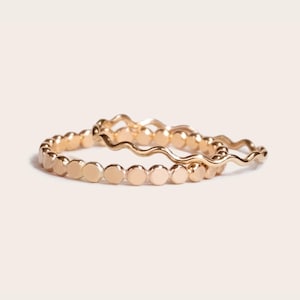 May include: Two gold rings. One ring features a series of small, flat circles, while the other has a wavy design. The rings are displayed on a light background, showcasing their shiny, metallic appearance. The rings are likely made of gold.
