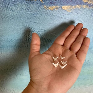 Puede incluir: Una mano sosteniendo seis grullas de papel origami blancas. Las grullas están dobladas en un estilo japonés tradicional.