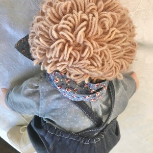 Puede incluir: Una mu&ntilde;eca hecha a mano con una camisa de estampado azul y blanco, un mono vaquero y una peluca de lana marr&oacute;n.