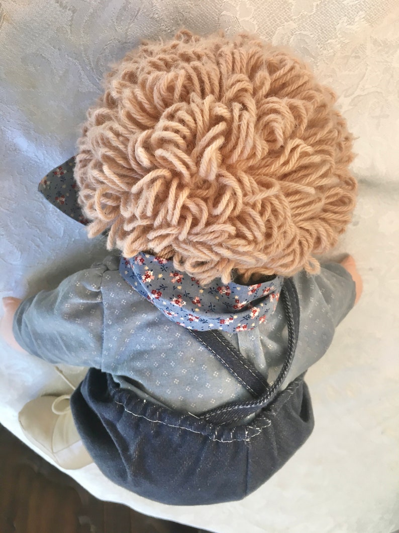 Puede incluir: Una mu&ntilde;eca hecha a mano con una camisa de estampado azul y blanco, un mono vaquero y una peluca de lana marr&oacute;n.