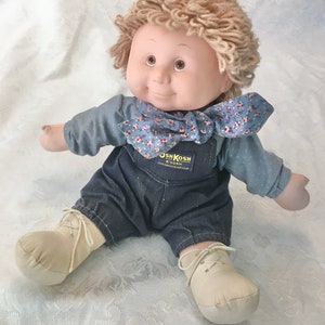 Puede incluir: Una mu&ntilde;eca de peluche con una camisa azul, un mono vaquero y una bufanda floral. La mu&ntilde;eca tiene el pelo de lana rubio y ojos marrones. La mu&ntilde;eca lleva zapatos blancos y est&aacute; sentada sobre una superficie blanca. El mono vaquero de la mu&ntilde;eca tiene el logotipo de OshKosh B'Gosh.