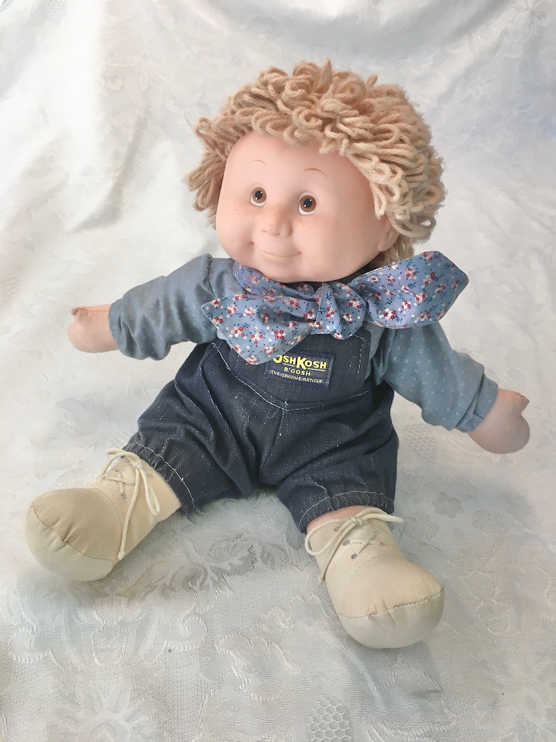 Puede incluir: Una mu&ntilde;eca de peluche con una camisa azul, un mono vaquero y una bufanda floral. La mu&ntilde;eca tiene el pelo de lana rubio y ojos marrones. La mu&ntilde;eca lleva zapatos blancos y est&aacute; sentada sobre una superficie blanca. El mono vaquero de la mu&ntilde;eca tiene el logotipo de OshKosh B'Gosh.