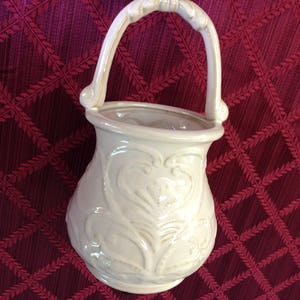 May include: White ceramic planter with a handle in the shape of a basket. The planter has an ornate design with raised details.